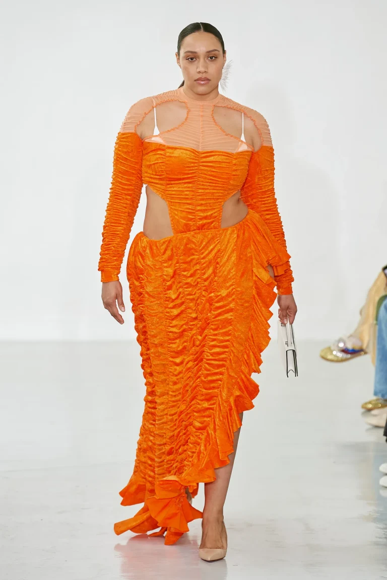 Une mannequin grande taille défile avec élégance et détermination, vêtue d’une robe orange éclatante à la coupe audacieuse. La robe présente un jeu de fronces texturées qui mettent en valeur les courbes, avec des découpes latérales assumées au niveau de la taille pour une allure moderne et sensuelle. Le haut de la tenue mêle transparence subtile et matières structurées, créant un effet graphique saisissant qui sublime le décolleté. Elle complète son look avec des escarpins nude et une pochette minimaliste, laissant la pièce maîtresse rayonner. Une silhouette vibrante qui incarne l'audace, la couleur et la célébration des corps magnifiés par l'identité GROSSES.