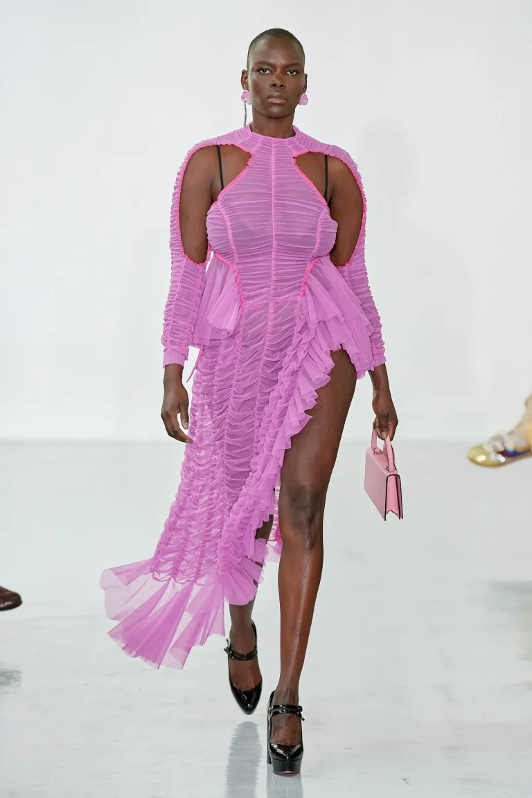 Une mannequin défile avec charisme dans une sublime robe rose translucide à effet plissé. La silhouette est structurée par des lignes graphiques et des volants asymétriques qui courent le long de la tenue, révélant subtilement la jambe avec une fente audacieuse. Les découpes dynamiques au niveau des épaules et du décolleté ajoutent une touche moderne et sensuelle. Elle complète le look avec des accessoires assortis : une petite pochette rose élégante et des escarpins noirs vernis, soulignant son allure affirmée. Cette tenue incarne parfaitement l’audace, la féminité et le glamour assumé propre à l’univers inclusif et fashion de GROSSES.