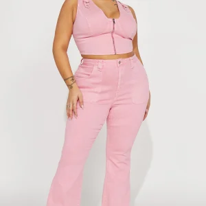 Une femme porte le top court rose nommé Nancy, fabriqué en denim stretch. Ce haut ajusté présente une fermeture éclair frontale, des bretelles larges et des détails de surpiqûres, créant un look moderne et flatteur. Elle l’associe à un pantalon en denim rose assorti à jambes évasées et des talons argentés, pour une tenue chic et coordonnée.