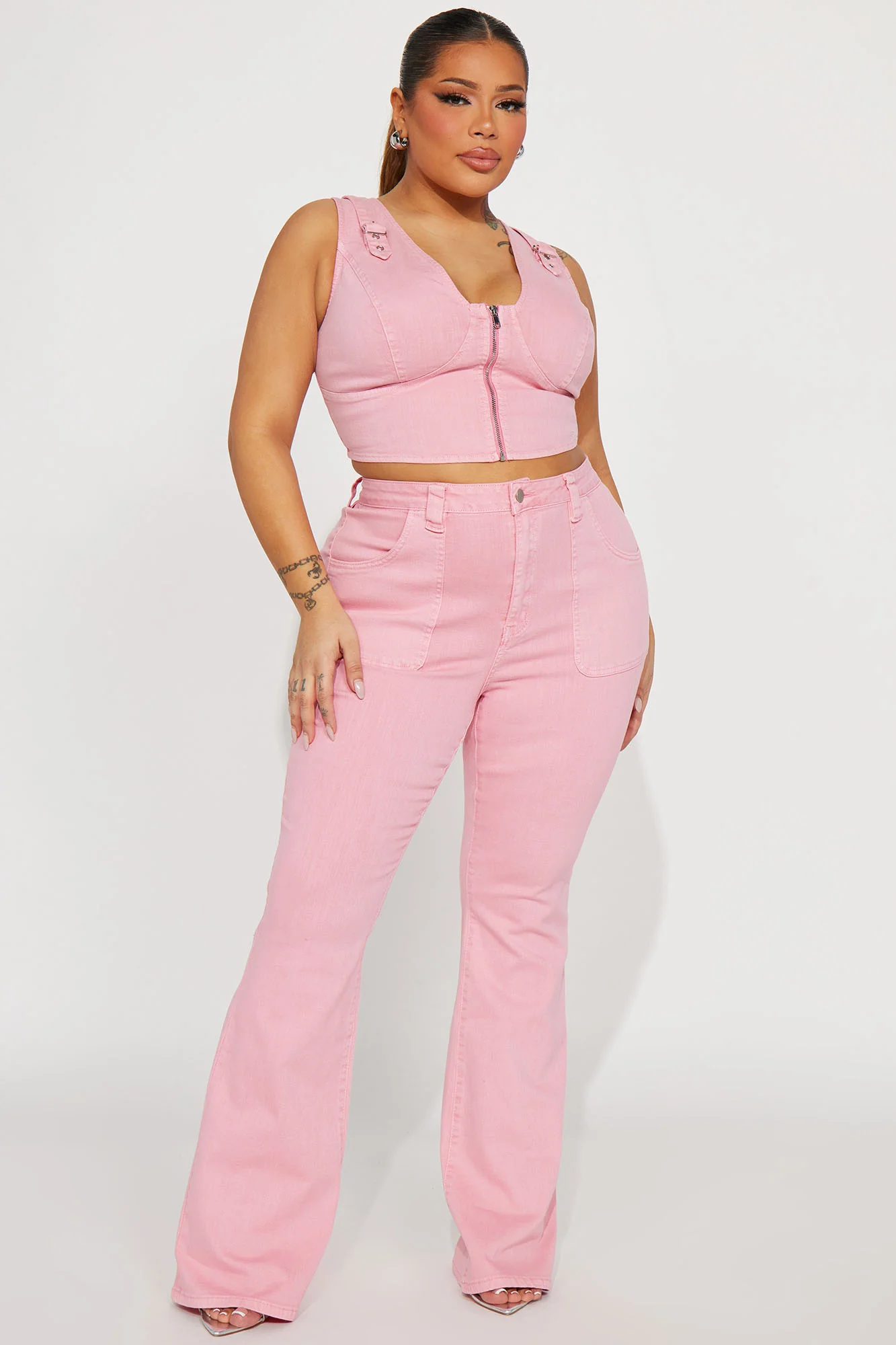 Une femme porte le top court rose nommé Nancy, fabriqué en denim stretch. Ce haut ajusté présente une fermeture éclair frontale, des bretelles larges et des détails de surpiqûres, créant un look moderne et flatteur. Elle l’associe à un pantalon en denim rose assorti à jambes évasées et des talons argentés, pour une tenue chic et coordonnée.