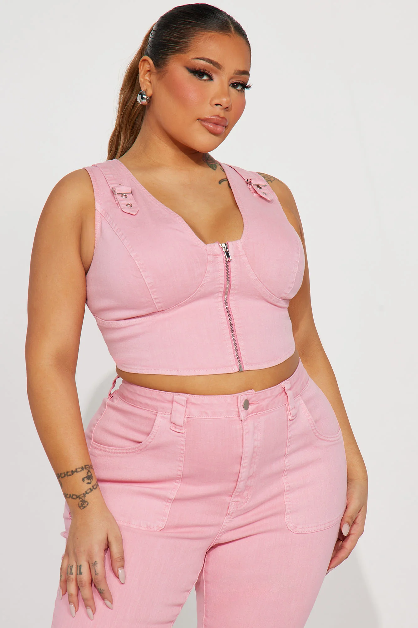 Une femme porte le top court rose nommé Nancy, fabriqué en denim stretch. Ce haut ajusté présente une fermeture éclair frontale, des bretelles larges et des détails de surpiqûres, créant un look moderne et flatteur. Elle l’associe à un pantalon en denim rose assorti à jambes évasées et des talons argentés, pour une tenue chic et coordonnée.