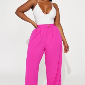 Une femme porte le pantalon fuchsia nommé Maya, confectionné en crêpe de soie mélangé (70 % soie et 30 % laine vierge). Ce pantalon à taille haute est accentué par un bouton doré et des plis marqués à l’avant, offrant une allure sophistiquée. Elle l’associe à un body blanc ajusté et des talons crème pour un look chic et audacieux.