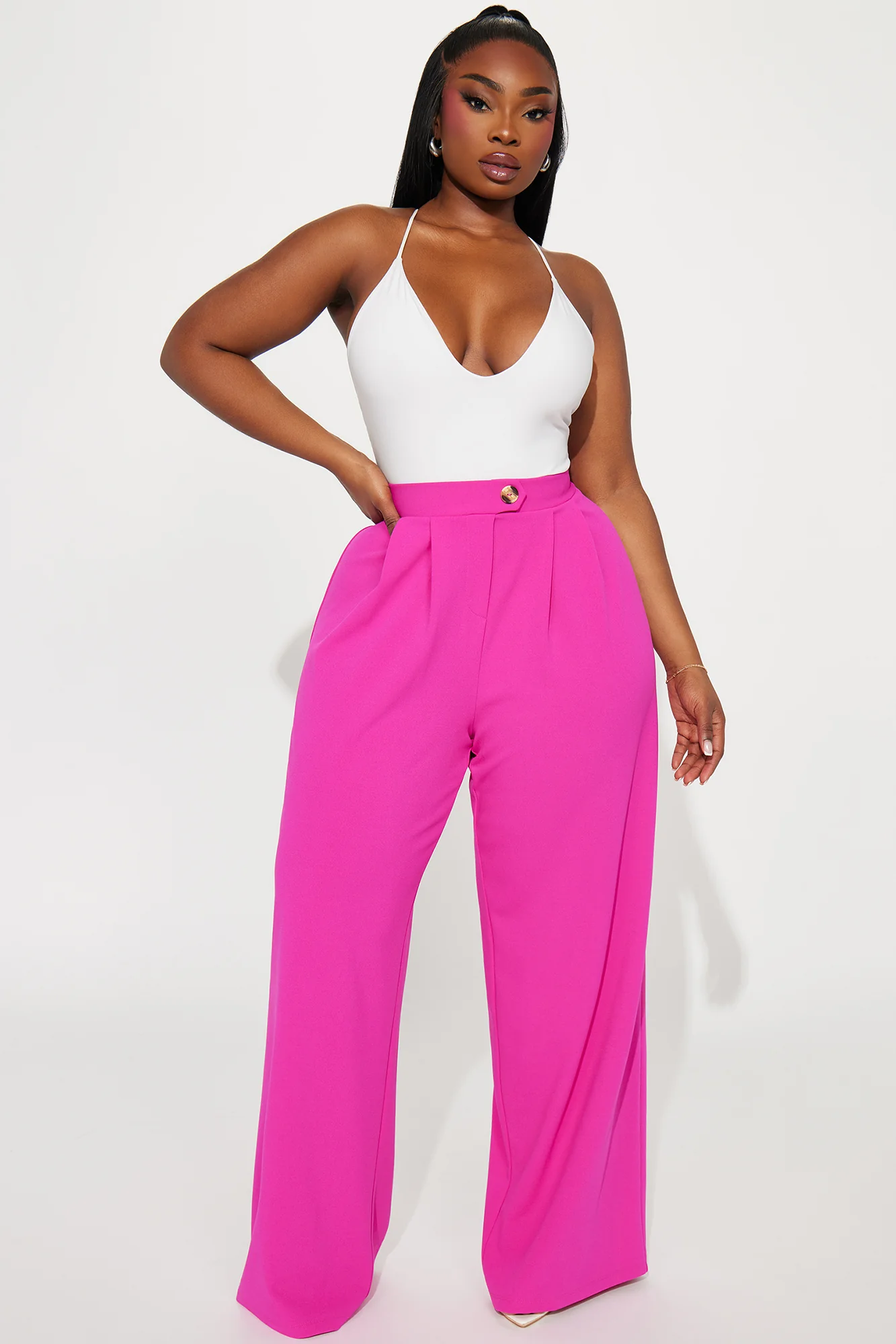 Une femme porte le pantalon fuchsia nommé Maya, confectionné en crêpe de soie mélangé (70 % soie et 30 % laine vierge). Ce pantalon à taille haute est accentué par un bouton doré et des plis marqués à l’avant, offrant une allure sophistiquée. Elle l’associe à un body blanc ajusté et des talons crème pour un look chic et audacieux.