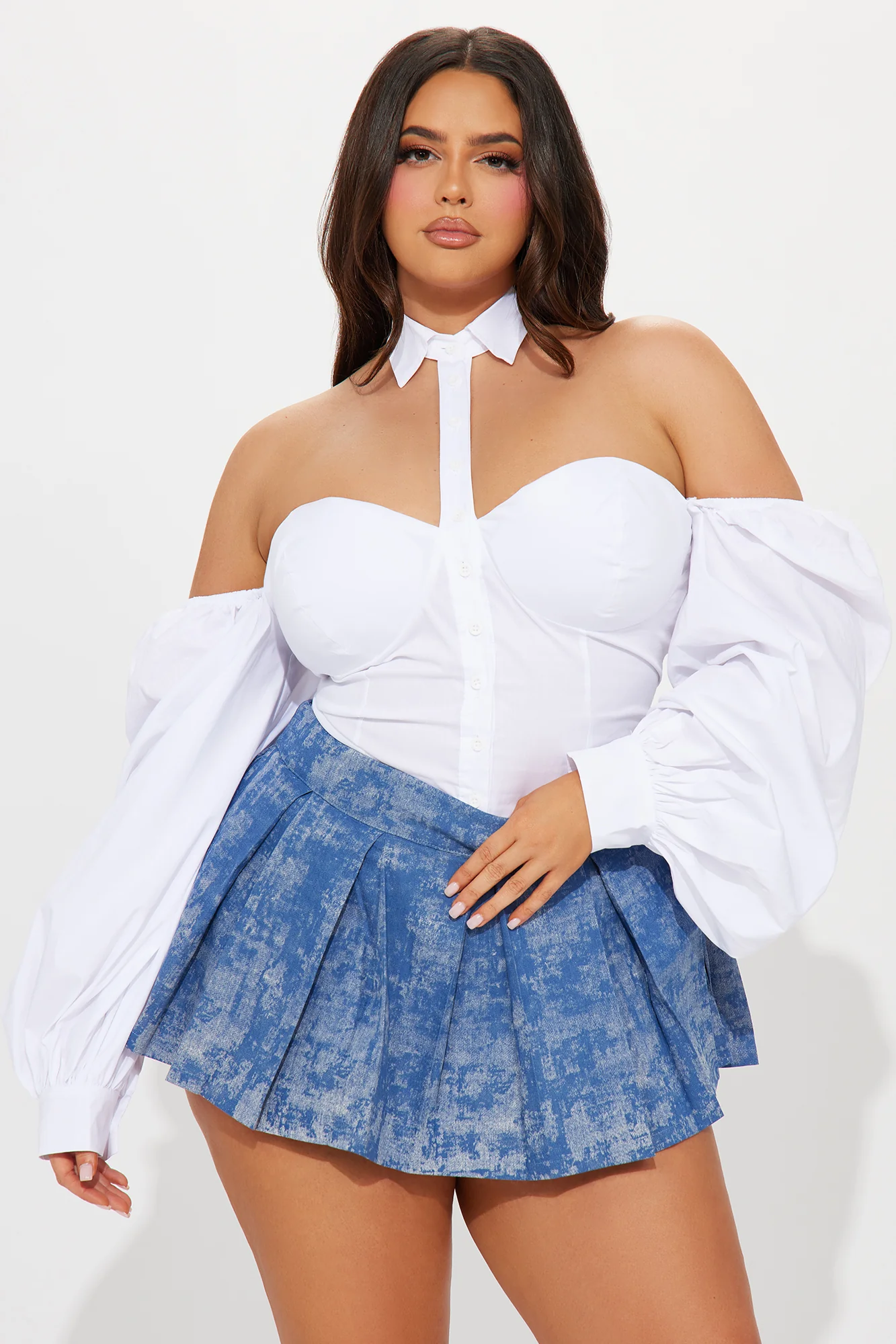 Une femme porte le body blanc nommé Cléo, confectionné en popeline de coton. Ce body associe un design bustier qui met en valeur le buste, un col chemise structuré et des manches longues volumineuses qui tombent sur les épaules, ajoutant une touche dramatique. Elle le combine avec une jupe plissée bleue, créant un look chic et moderne.