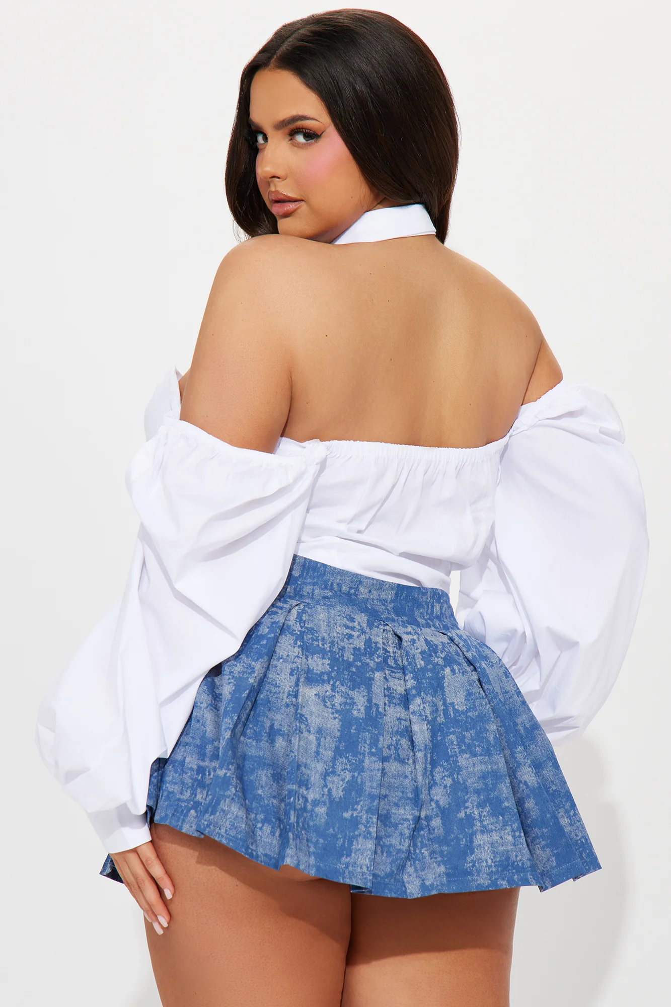 Une femme porte le body blanc nommé Cléo, confectionné en popeline de coton. Ce body associe un design bustier qui met en valeur le buste, un col chemise structuré et des manches longues volumineuses qui tombent sur les épaules, ajoutant une touche dramatique. Elle le combine avec une jupe plissée bleue, créant un look chic et moderne.