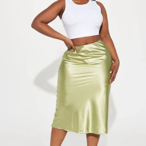 Une femme porte la jupe midi verte nommée Coralie, fabriquée en satin de soie. La jupe fluide, légèrement évasée, suit les mouvements avec élégance. Elle est associée à un haut blanc ajusté et des talons transparents, créant un look minimaliste et raffiné.