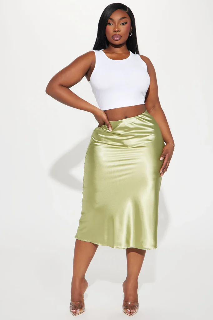 Une femme porte la jupe midi verte nommée Coralie, fabriquée en satin de soie. La jupe fluide, légèrement évasée, suit les mouvements avec élégance. Elle est associée à un haut blanc ajusté et des talons transparents, créant un look minimaliste et raffiné.