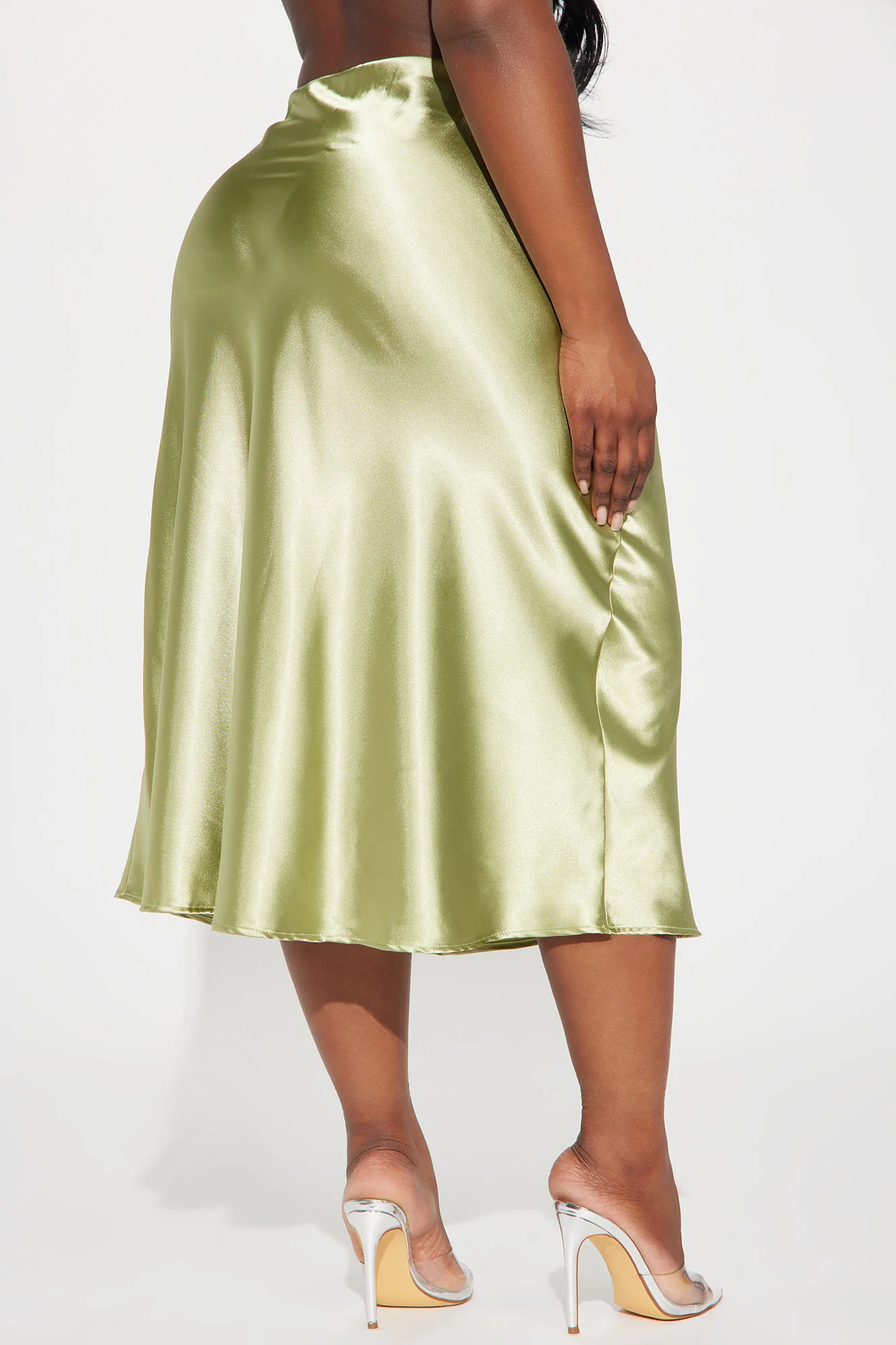 Une femme porte la jupe midi verte nommée Coralie, fabriquée en satin de soie. La jupe fluide, légèrement évasée, suit les mouvements avec élégance. Elle est associée à des talons transparents, créant un look minimaliste et raffiné.