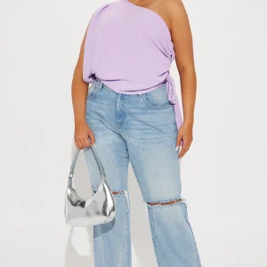 Une femme porte le top asymétrique lavande nommé Alek, confectionné en jersey de coton biologique. Le haut dévoile une épaule avec une coupe fluide et des fronces ajustables sur le côté, attachées par une ceinture. Elle l'associe à un jean large et déchiré bleu clair, des sandales à talons argentées et un sac à main métallisé argent, créant un look moderne, lumineux et décontracté.
