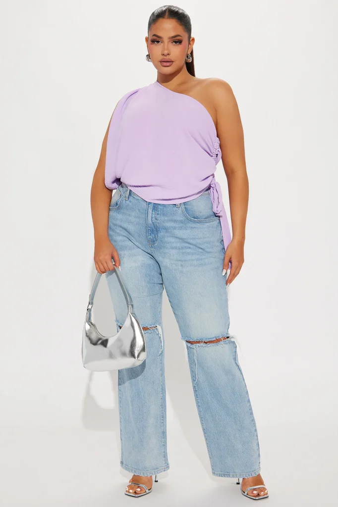 Une femme porte le top asymétrique lavande nommé Alek, confectionné en jersey de coton biologique. Le haut dévoile une épaule avec une coupe fluide et des fronces ajustables sur le côté, attachées par une ceinture. Elle l'associe à un jean large et déchiré bleu clair, des sandales à talons argentées et un sac à main métallisé argent, créant un look moderne, lumineux et décontracté.