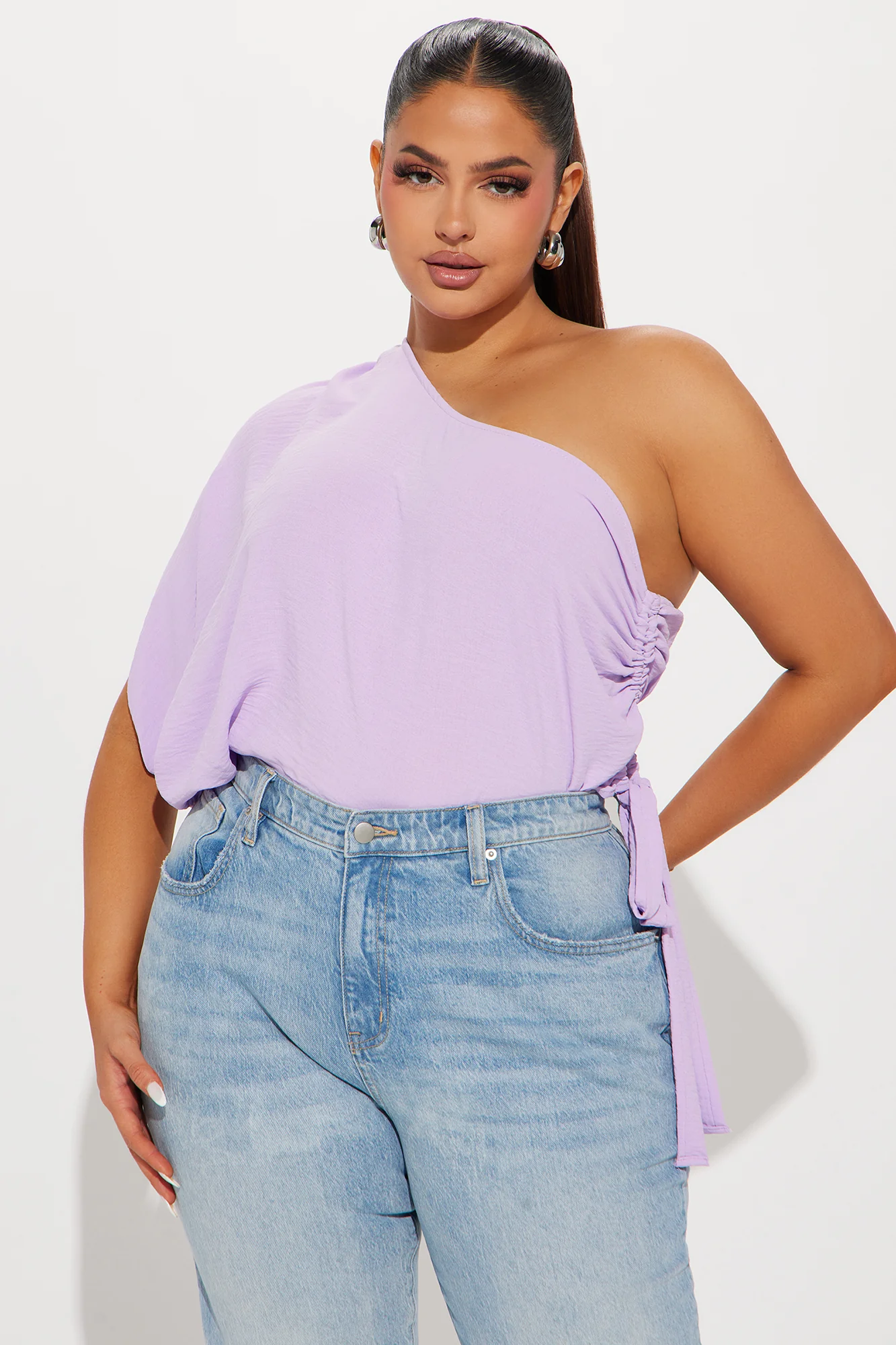 Une femme porte le top asymétrique lavande nommé Alek, fabriqué en jersey de coton biologique. Le haut dévoile une épaule avec une coupe fluide et légère, tandis que des fronces sur le côté, accompagnées d’une ceinture ajustable, créent une silhouette personnalisée. Elle l’associe à un jean bleu clair pour un look décontracté et moderne.