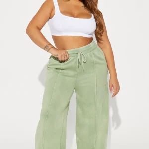 Une femme porte le pantalon large vert nommé Julie, confectionné en molleton de coton. Ce pantalon fluide, doté de coutures apparentes et d’une taille élastiquée avec cordon, est associé à un crop top blanc et des baskets, créant un look décontracté et moderne.