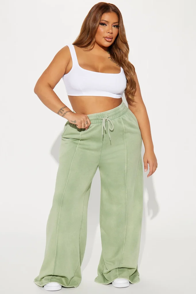 Une femme porte le pantalon large vert nommé Julie, confectionné en molleton de coton. Ce pantalon fluide, doté de coutures apparentes et d’une taille élastiquée avec cordon, est associé à un crop top blanc et des baskets, créant un look décontracté et moderne.