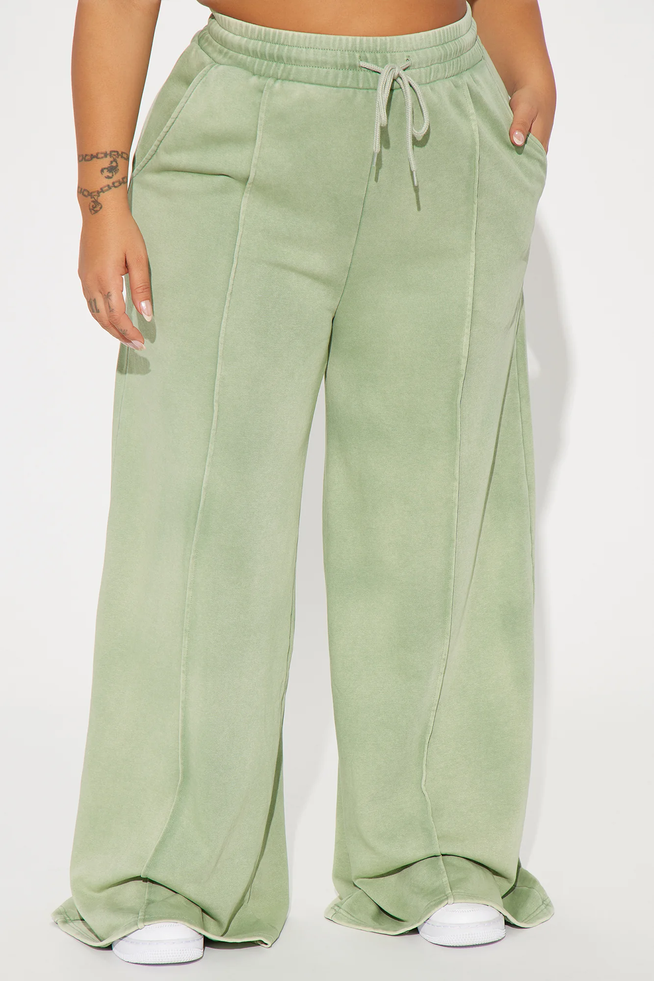 Une femme porte le pantalon large vert nommé Julie, confectionné en molleton de coton. Ce pantalon fluide, doté de coutures apparentes et d’une taille élastiquée avec cordon, est associé à des baskets blanches, créant un look décontracté et moderne.