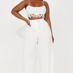 Une femme porte le pantalon blanc nommé Sandra, confectionné en crêpe de laine. Ce pantalon à taille haute, orné d’un bouton doré et doté d’une coupe large, est associé à un crop top en dentelle blanche, créant un look sophistiqué et féminin.