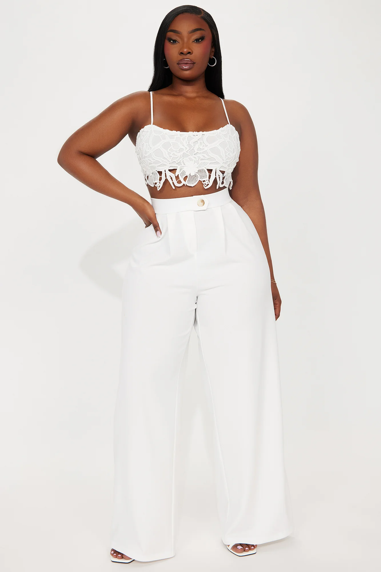 Une femme porte le pantalon blanc nommé Sandra, confectionné en crêpe de laine. Ce pantalon à taille haute, orné d’un bouton doré et doté d’une coupe large, est associé à un crop top en dentelle blanche, créant un look sophistiqué et féminin.