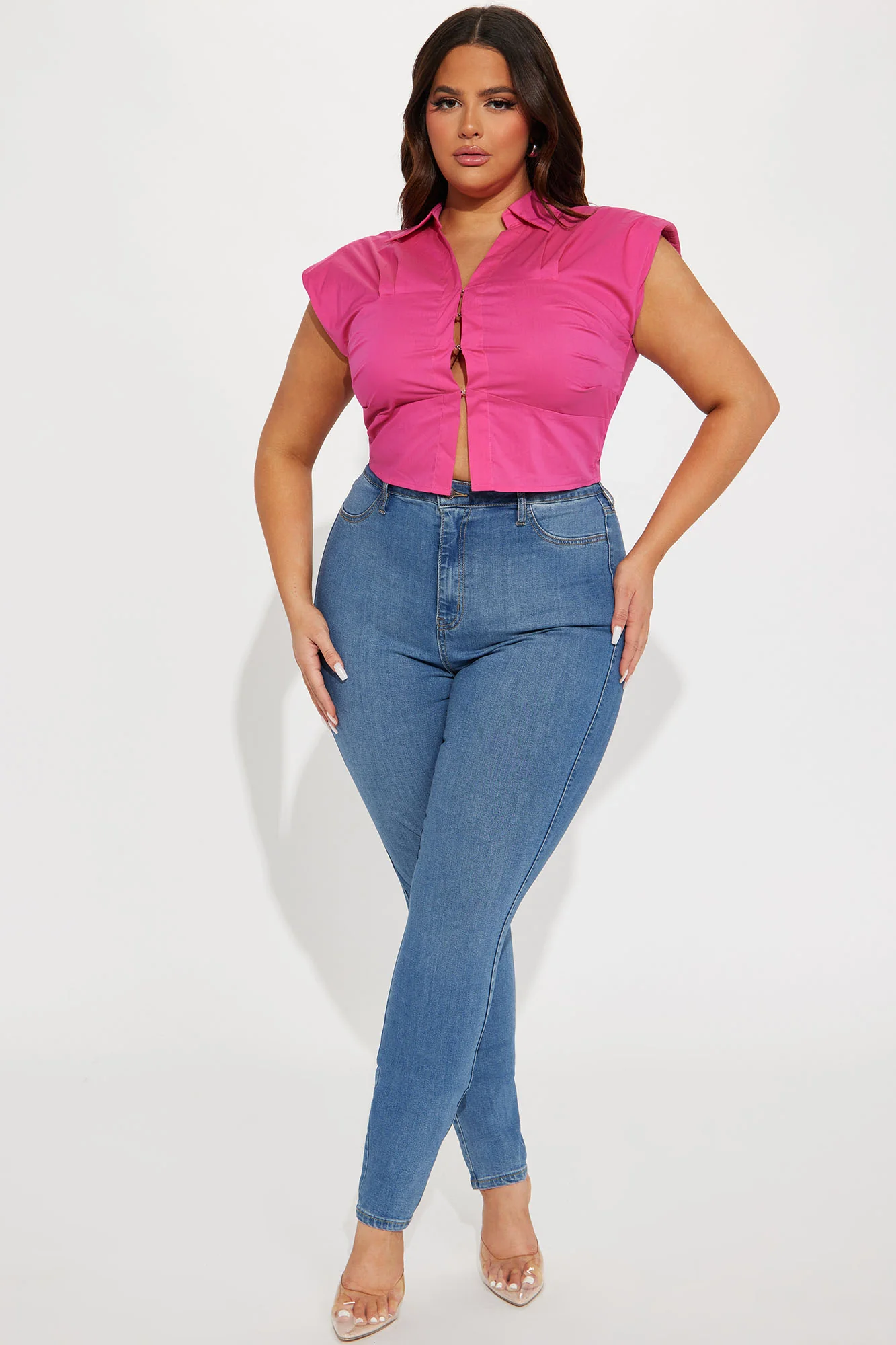 Une femme curvy porte un top rose vif en coton satiné, avec un col chemise structuré et une coupe ajustée qui met en valeur sa silhouette. Le top est légèrement ouvert à l'avant pour une touche sensuelle. Elle l'associe à un jean skinny bleu et des talons transparents, créant un look moderne et chic.