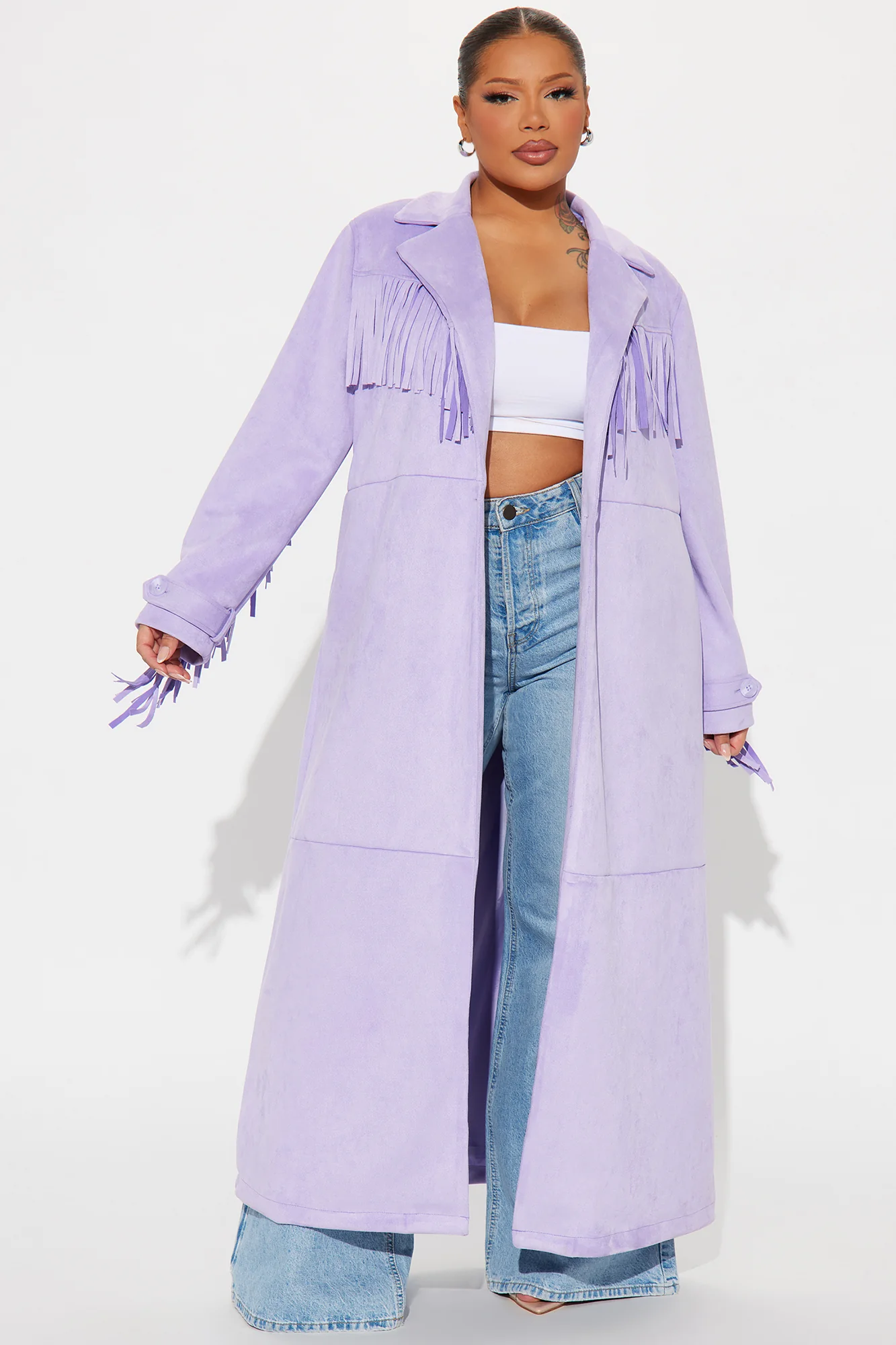 Une femme porte le manteau long lavande nommé Capucine, confectionné en suédine de fibres naturelles. Ce manteau fluide, orné de franges sur les épaules et les manches, est associé à un jean évasé et un crop top blanc, créant un look bohème et moderne.