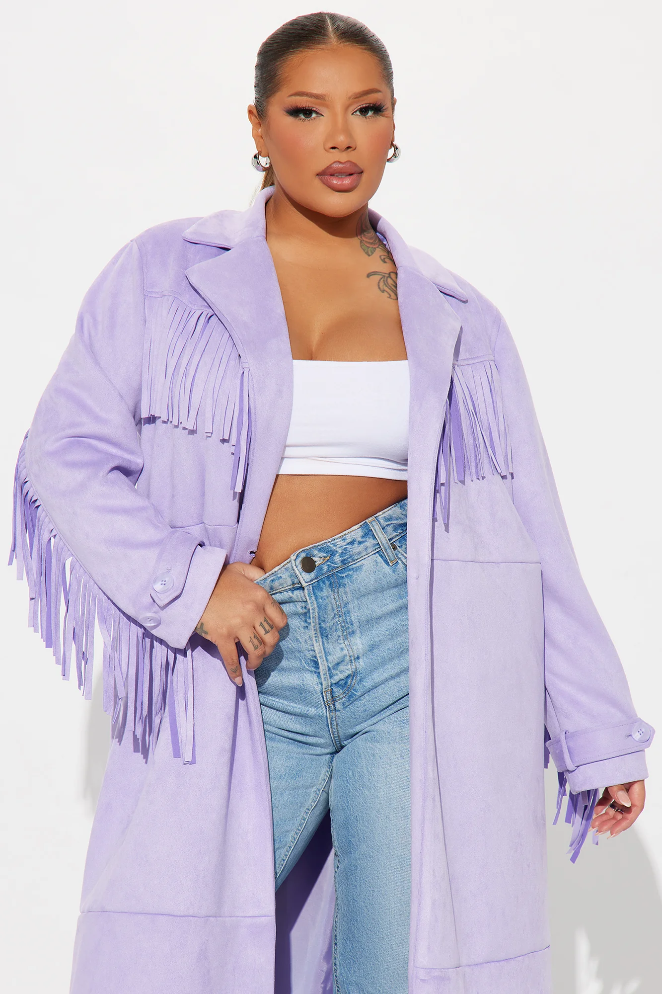Une femme porte le manteau long lavande nommé Capucine, confectionné en suédine de fibres naturelles. Ce manteau fluide, orné de franges sur les épaules et les manches, est associé à un jean évasé et un crop top blanc, créant un look bohème et moderne.