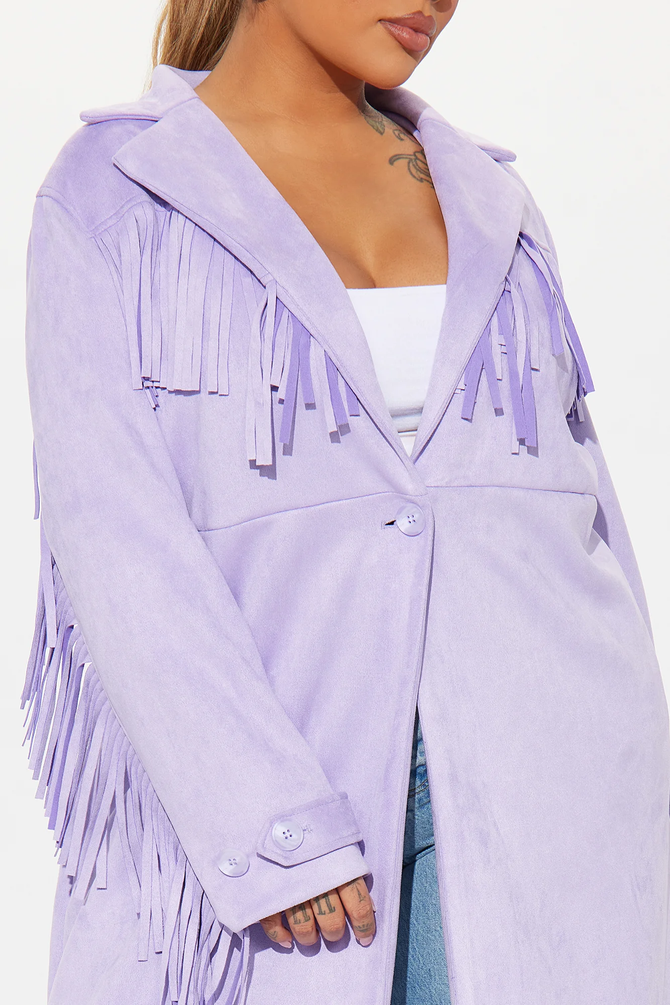 Une femme porte le manteau long lavande nommé Capucine, confectionné en suédine de fibres naturelles. Ce manteau fluide, orné de franges sur les épaules et les manches, est associé à un jean évasé et un crop top blanc, créant un look bohème et moderne.