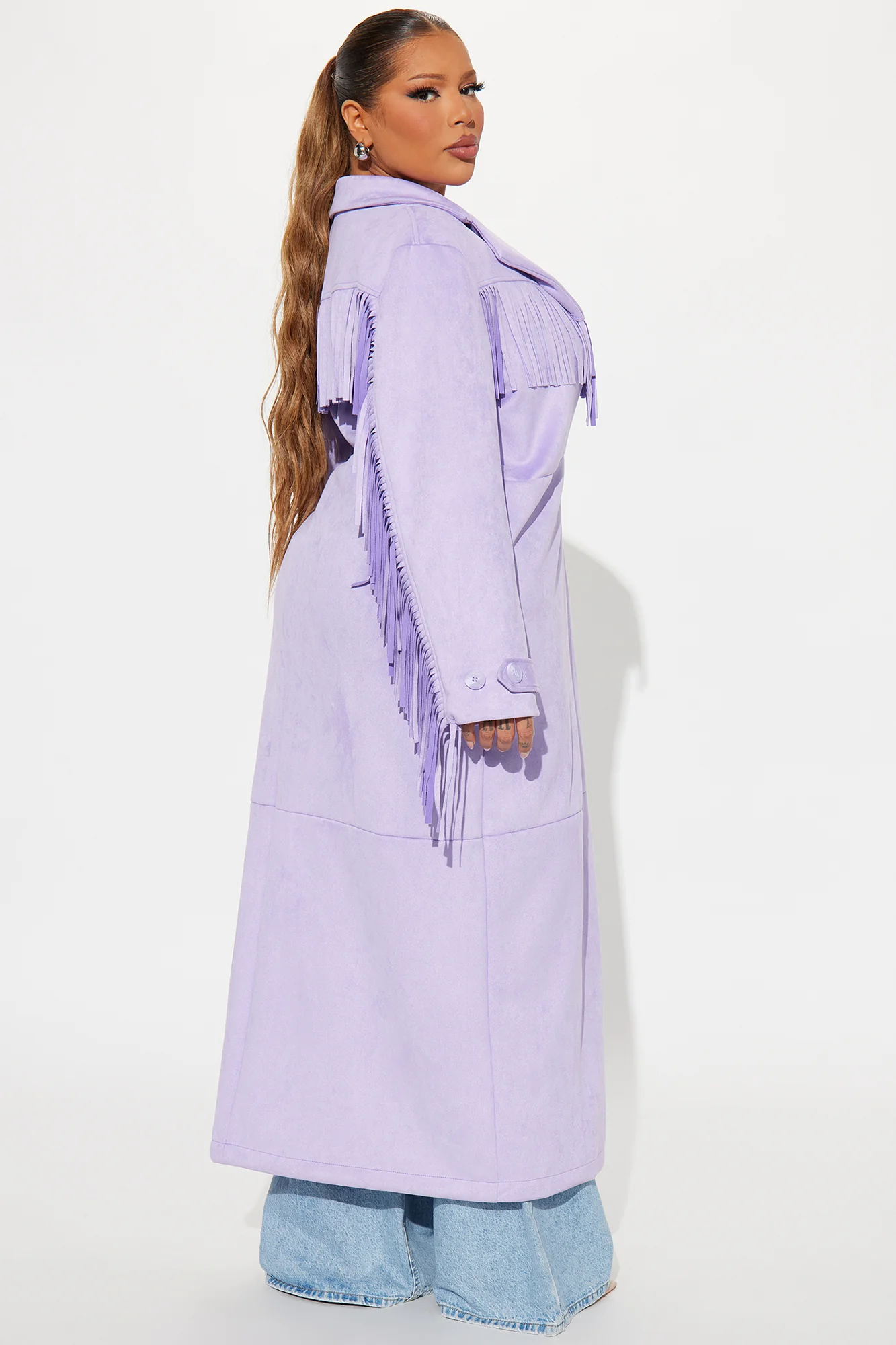 Une femme porte le manteau long lavande nommé Capucine, confectionné en suédine de fibres naturelles. Ce manteau fluide, orné de franges sur les épaules et les manches, est associé à un jean évasé et un crop top blanc, créant un look bohème et moderne.