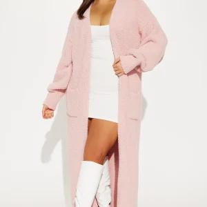 Une femme porte le gilet long rose nommé Jacqueline, confectionné en maille bouclée de laine mérinos et cachemire. Ce gilet oversize, doté de poches et d’une coupe fluide, est associé à une robe blanche ajustée et des bottes blanches, créant un look féminin et cosy.