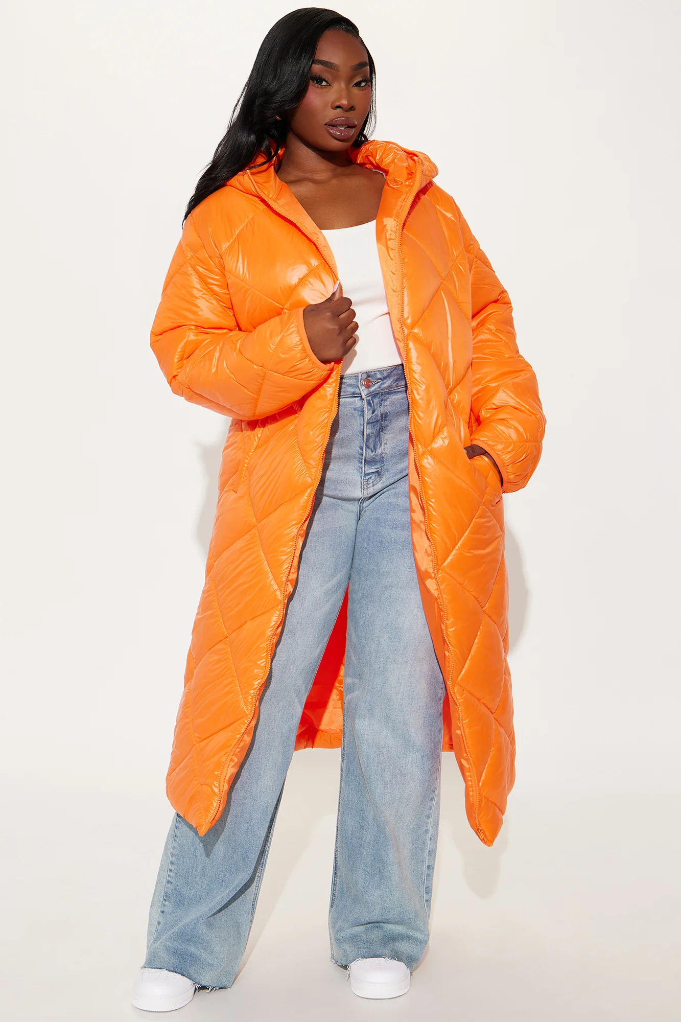 Une femme porte la doudoune longue orange nommée Caroline, confectionnée en tissu matelassé imperméable à base de fibres recyclées. Cette doudoune oversize, dotée d’une capuche et d’une fermeture zippée, est associée à un jean et un top blanc, pour un look hivernal urbain et audacieux.
