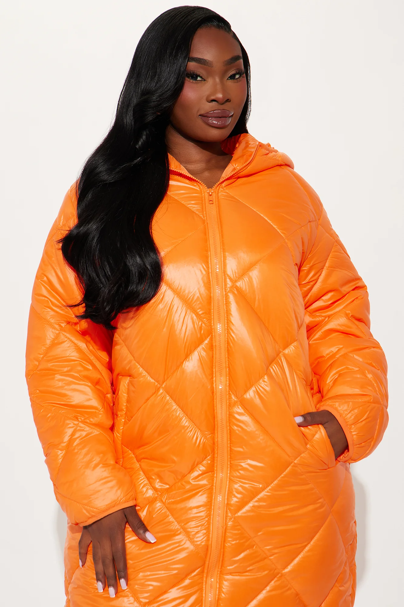 Une femme porte la doudoune longue orange nommée Caroline, confectionnée en tissu matelassé imperméable à base de fibres recyclées. Cette doudoune oversize, dotée d’une capuche et d’une fermeture zippée, est associée à un jean et un top blanc, pour un look hivernal urbain et audacieux.