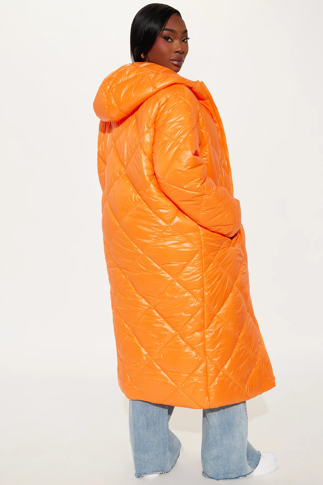 Une femme porte la doudoune longue orange nommée Caroline, confectionnée en tissu matelassé imperméable à base de fibres recyclées. Cette doudoune oversize, dotée d’une capuche et d’une fermeture zippée, est associée à un jean et un top blanc, pour un look hivernal urbain et audacieux.