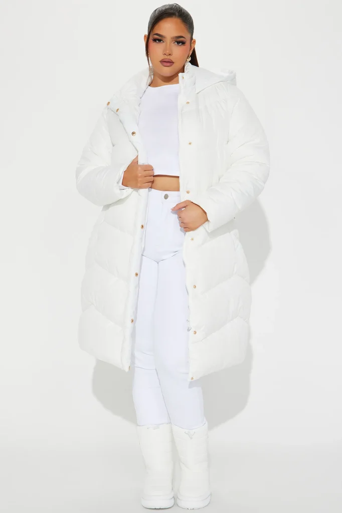 Une femme porte la doudoune blanche nommée Billie, confectionnée en tissu matelassé à base de fibres de maïs Sorona. Cette pièce mi-longue et ajustée, dotée d’une capuche amovible et de boutons dorés, est associée à un ensemble blanc et des bottes, créant un look hivernal chic et moderne.