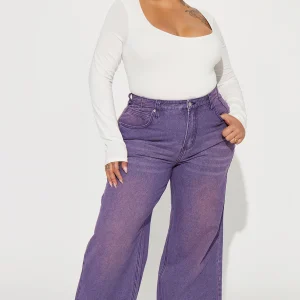 Une femme porte le pantalon violet nommé Manon, confectionné en denim de coton. Ce pantalon à jambes larges et taille haute est doté d’ourlets effilés, associé à un body blanc ajusté et des baskets, créant un look décontracté et moderne.
