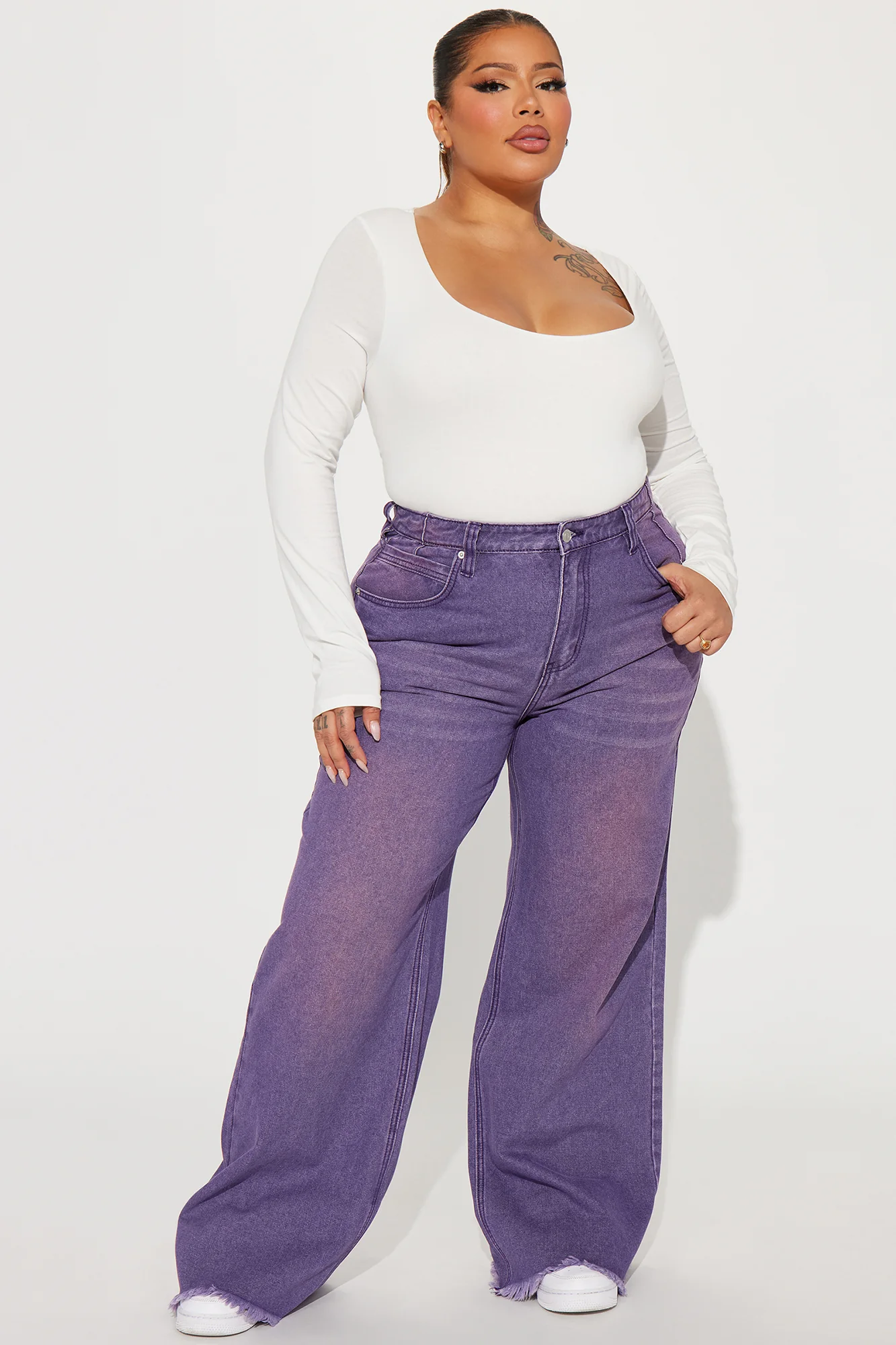 Une femme porte le pantalon violet nommé Manon, confectionné en denim de coton. Ce pantalon à jambes larges et taille haute est doté d’ourlets effilés, associé à un body blanc ajusté et des baskets, créant un look décontracté et moderne.