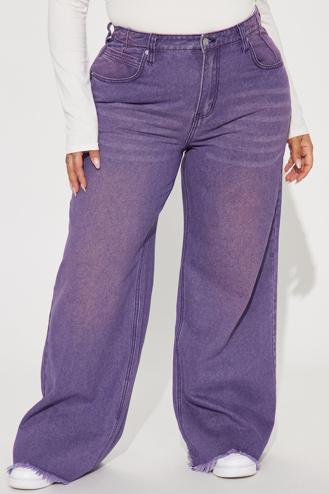 Une femme porte le pantalon violet nommé Manon, confectionné en denim de coton. Ce pantalon à jambes larges et taille haute est doté d’ourlets effilés, associé à un body blanc ajusté et des baskets, créant un look décontracté et moderne.