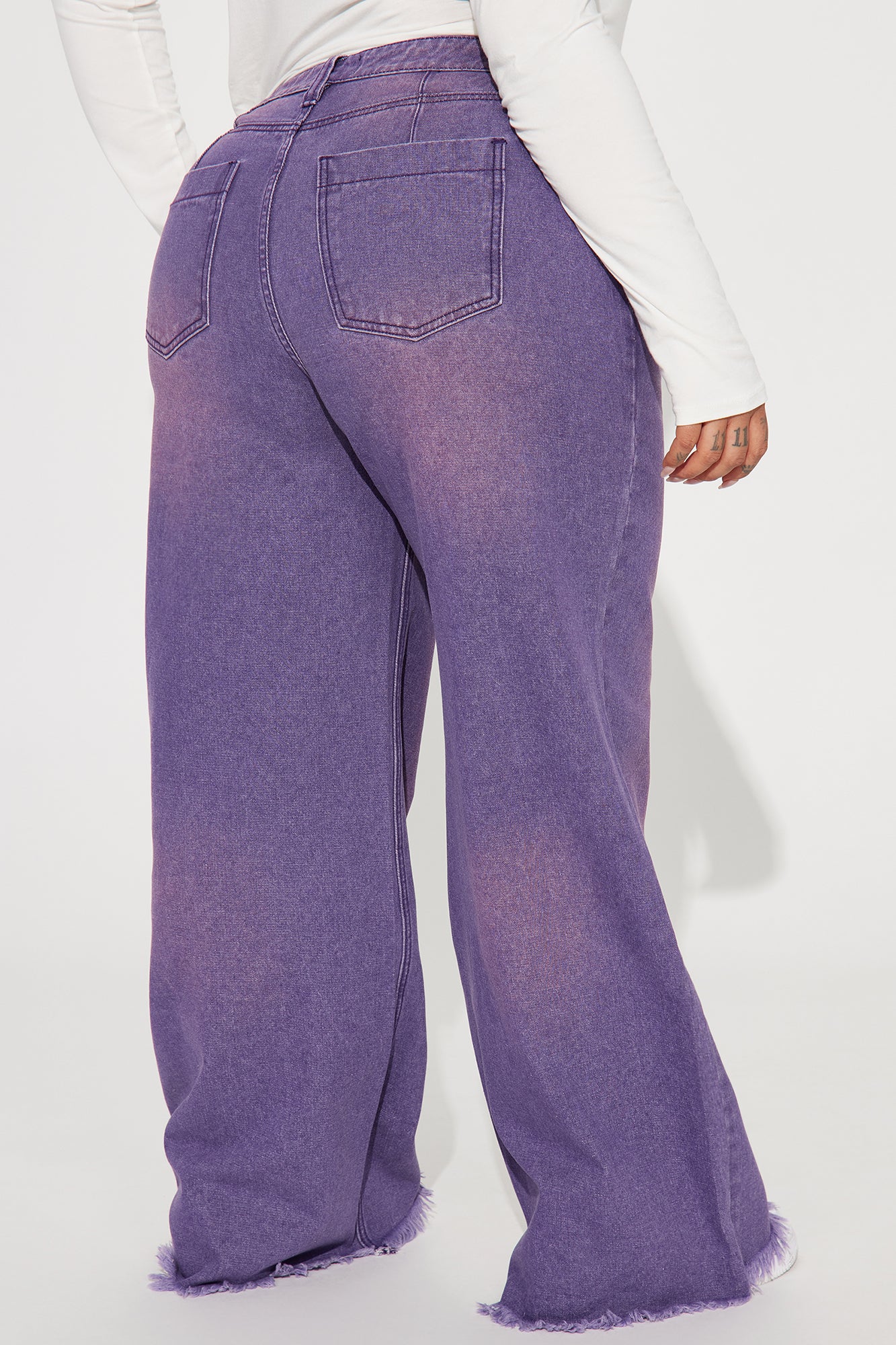 Une femme porte le pantalon violet nommé Manon, confectionné en denim de coton. Ce pantalon à jambes larges et taille haute est doté d’ourlets effilés, associé à un body blanc ajusté et des baskets, créant un look décontracté et moderne.