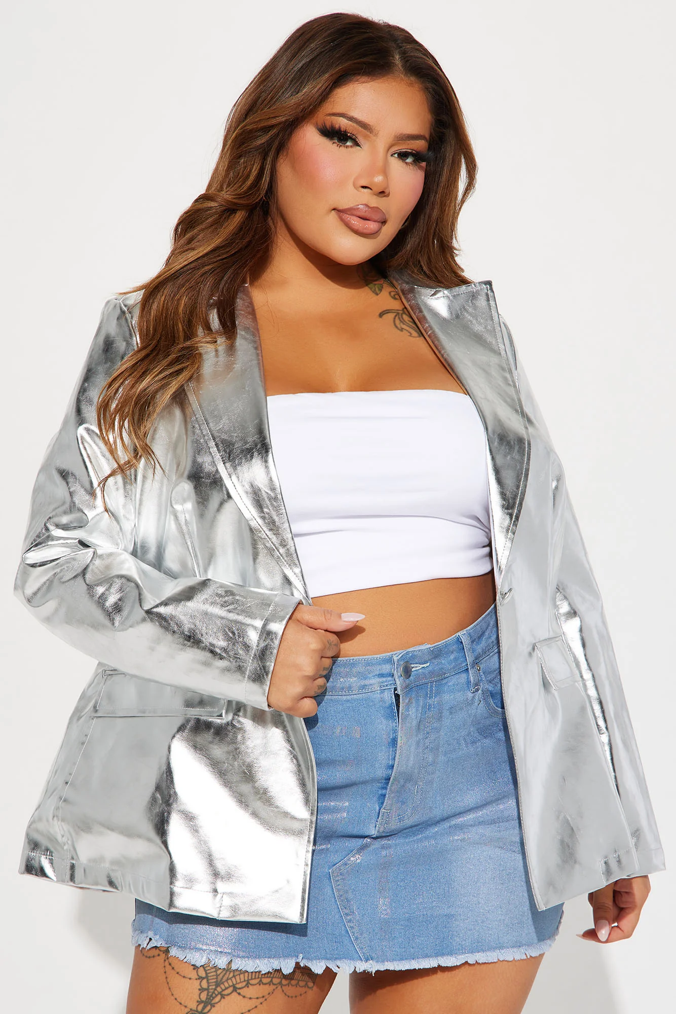 Une femme porte la veste argentée nommée Frida, confectionnée en cuir végétal métallisé. Ce blazer structuré, doté d’un fini brillant et de poches plaquées, est associé à un crop top blanc et une mini-jupe en denim, créant un look audacieux et contemporain.