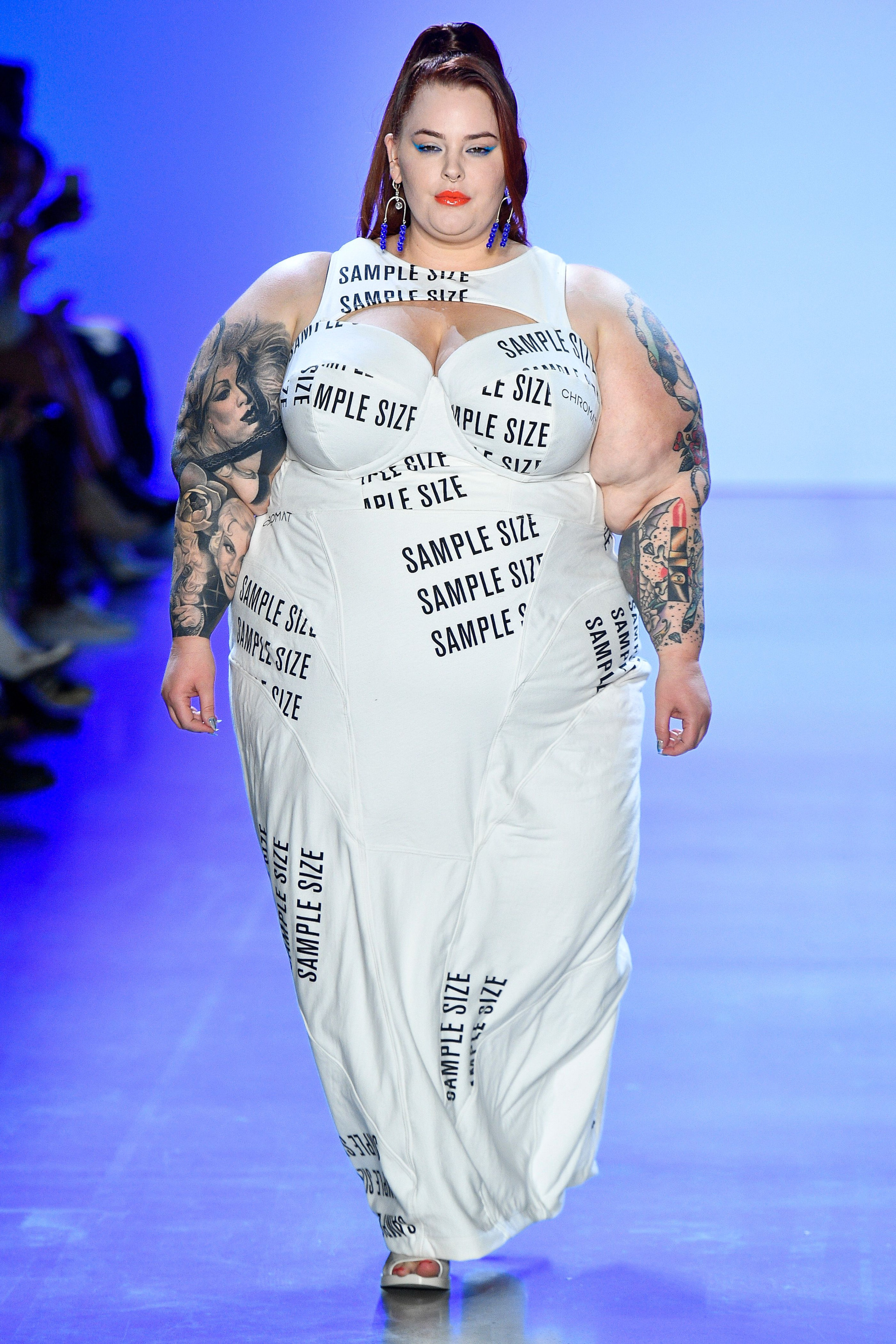 Tess Holliday défile en robe blanche imprimée «Sample Size» pour Chromat en 2019, tatouages visibles, attitude forte.