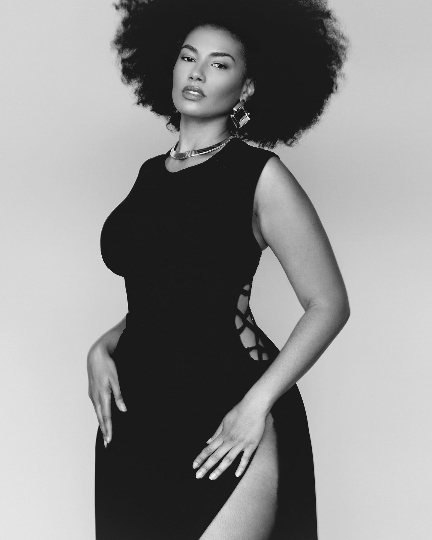 Shadia Ampadu pose fièrement en robe noire fendue, coiffure afro volumineuse, regard assuré, portrait noir et blanc élégant.
